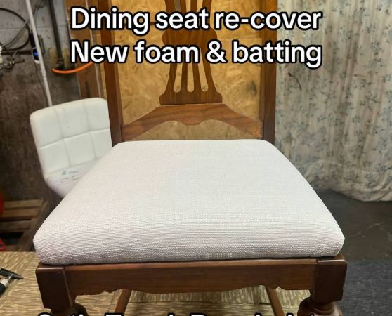 A wooden dining chair with a medium to dark-toned hardwood frame, featuring turned legs and a vertical slat backrest with a curved top rail, upholstered in white-colored fabric