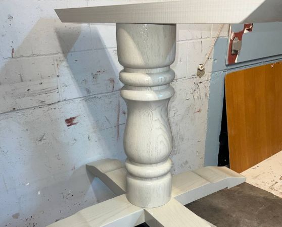 A partially assembled white wooden table base with a turned pedestal and cross supports.