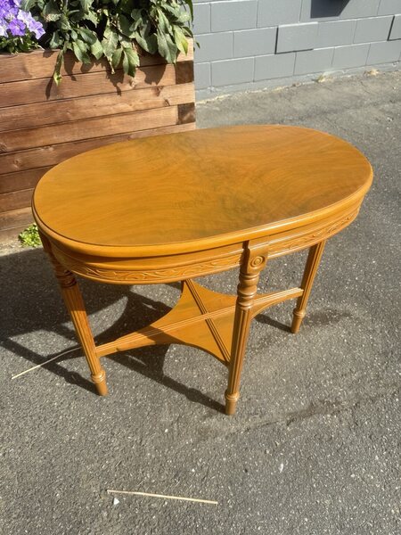Oval wooden accent table with a golden finish, carved apron, fluted legs and a shaped lower shelf, viewed from an angle.
