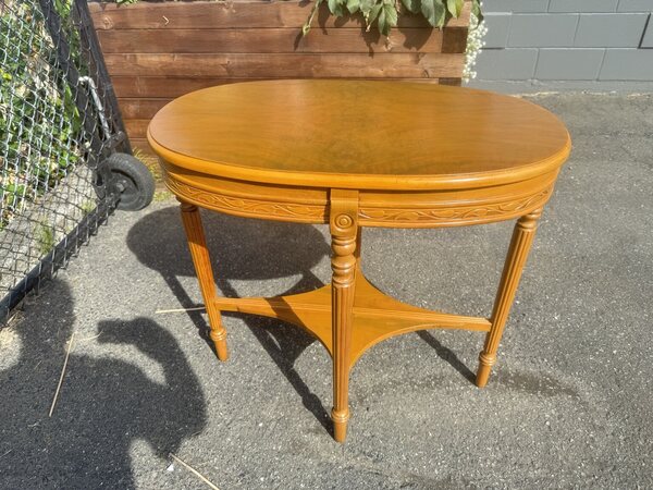 Front view of a golden-finished oval accent table with a decorative carved apron, fluted legs and lower shelf.