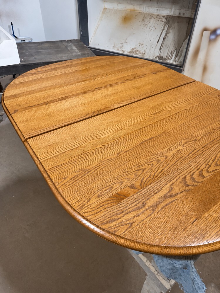 Large oval oak dining tabletop with a warm golden finish and visible grain, resting on padded supports in the Satin Touch workshop.