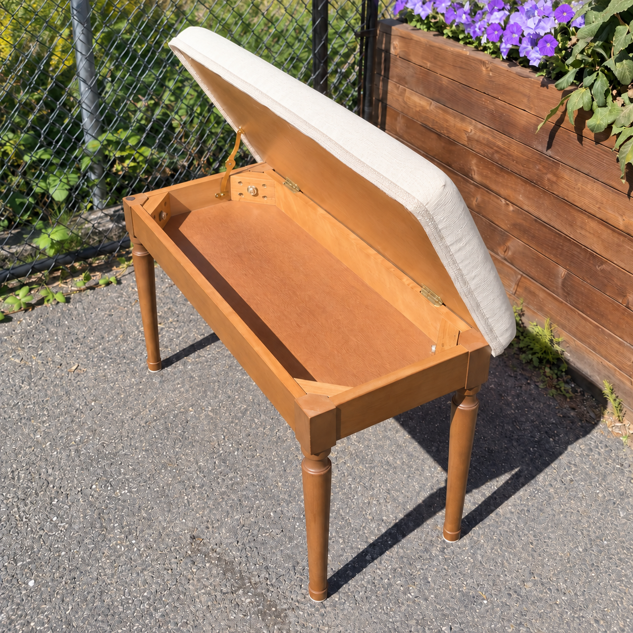 Open wooden piano bench showing its storage compartment and cream upholstered lid with decorative trim.